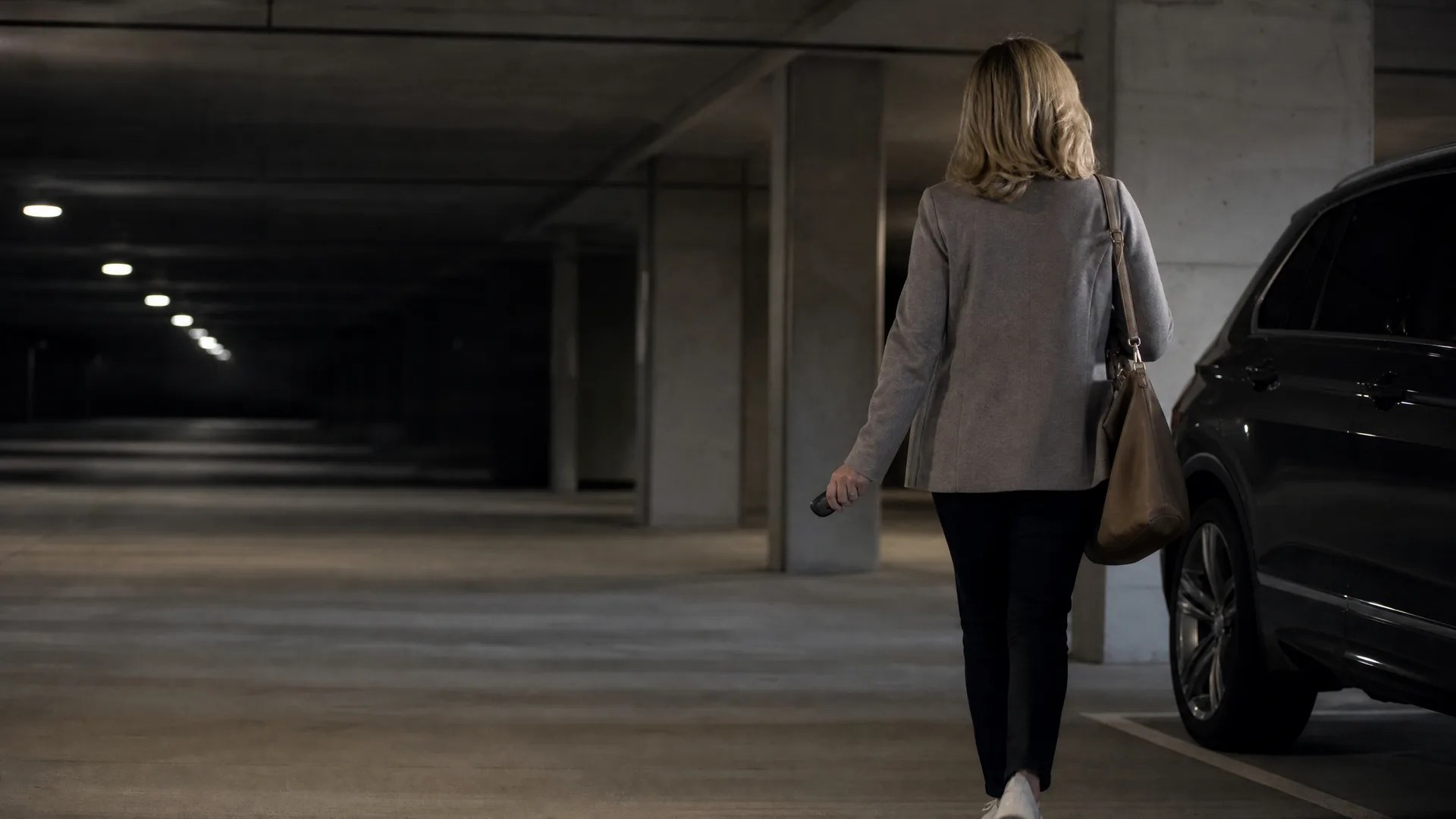 A woman in a grey coat and black pants, carrying a shoulder bag and holding a key fob, walks away from the camera toward a dark SUV in a dimly lit concrete parking garage.