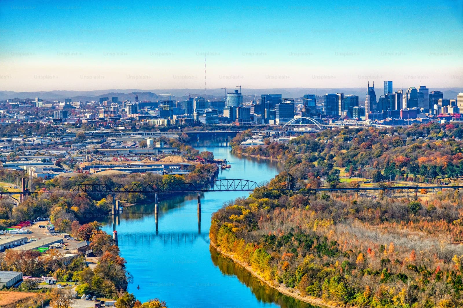 Aerial view of city skyline with river winding through autumn trees and multiple bridges under blue sky
